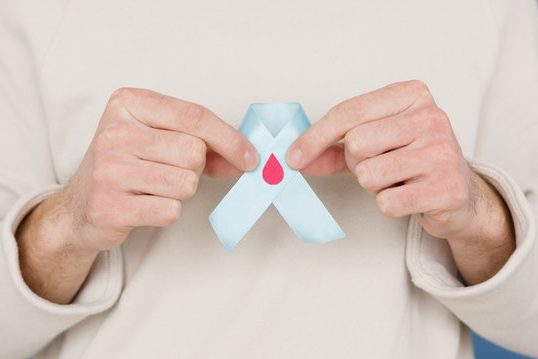 doacao-de-sangue Torso de homem segurando laço com a uma gota vermelha.